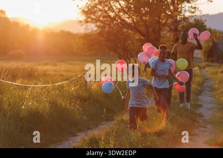 Ein alleinerziehender Vater und seine beiden Kinder laufen mit bunten Ballons auf einem Feld bei Sonnenuntergang. Sie teilen fröhliches Lachen und Lächeln. Stockfoto