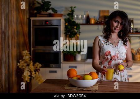 Frau in der Nähe von Orangen, die Frühstück vorbereitet, rustikale Küche mit Frau und frische Orangen auf der Theke, heimelige Frühstücksszene mit Frau mit Orangen in Stockfoto