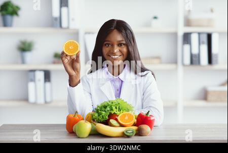 Der Ernährungsberater hält Orange, während er in Office Workspace von frischem Obst und Gemüse umgeben ist Stockfoto