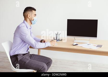 Der Mann trägt eine Maske und sitzt am Schreibtisch und schreibt Notizen während der Arbeit von zu Hause aus im Büro Stockfoto