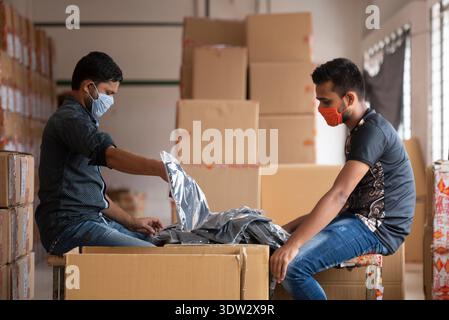 Professionelles Bild von zwei Arbeitern, die Gesichtsmasken tragen, die Kartonschachteln in einem gut beleuchteten Lager handhaben und Teamarbeit und Sicherheit während des Umzugs fördern Stockfoto