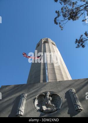 Ein flacher Blick auf den Coit Tower, der sich über seinem geformten Sockel erhebt, begleitet von einer amerikanischen Flagge. Das Wahrzeichen hebt sich deutlich vom Himmel ab. Stockfoto