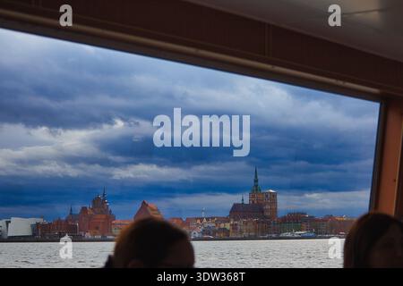 Touristen auf einem Ausflugsboot sehen die Stadtlandschaft von Stralsund mit der Marienkirche und dem Ozeaneum unter einem dramatischen, bewölkten Himmel Stockfoto