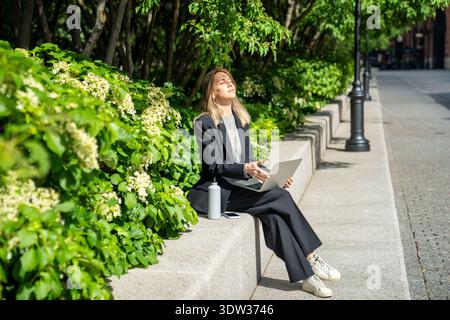 Entspannte Geschäftsfrau in der täglichen Pause im Park, die unter Bäumen sitzt, lächelt und blinzt in der Sonne. Stockfoto