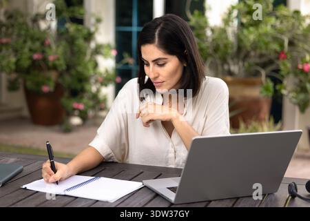 Fokussierte Erwachsene Studentin, die im Copybook am Laptop schreibt Stockfoto