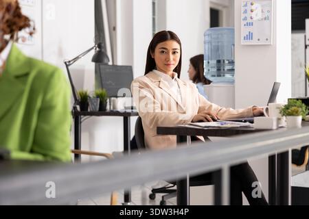 Eine selbstbewusste Führungskraft blickt im Unternehmensbüro fokussiert und bereit in die Kamera. Asiatische Geschäftsfrau saß am Schreibtisch und drückte ihr Engagement für den Erfolg der Marketingfirma aus. Stockfoto