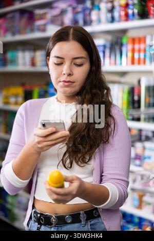 Porträt einer lächelnden Verbraucherin, die ein Produkt in der Parfümerie auswählt und zum Scannen des Barcodes ein Smartphone verwendet Stockfoto