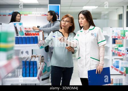 Medizinberater zeigt auf pharmazeutische Produkte in Regalen und gibt neugierigen Kunden in einer modernen Drogerie detaillierte Informationen. Asiatische Frauen, die sich Arzneimittelpakete im Apothekengang ansehen. Stockfoto
