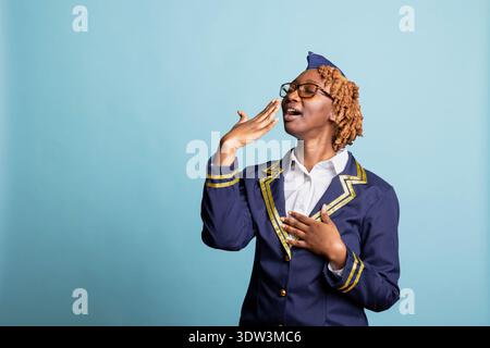 Müde Flugbegleiterin in Uniform Gähnen und Mund vor blauem Hintergrund abdecken. Schwarzlufthostess erlebt Ermüdung, lange Flüge und Erschöpfung in Luftfahrt- und Reiseberufen. Stockfoto