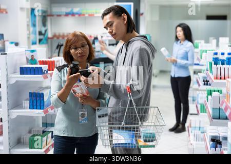 Asiatischer Mann zeigt dem Apothekenangestellten Smartphone und bittet um Hilfe bei der Suche nach Gesundheitsprodukten. Die weibliche Drogeriemärtnerin hält Medikamente und hilft männlichen Kunden basierend auf dem Bild auf dem Gerät. Stockfoto