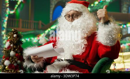 Der Weihnachtsmann liest Briefe von Kindern in gemütlichem Weihnachtsset, auf dem ein Schild mit den Daumen nach oben steht. Mann, der positive Handgesten macht, auf Wunschlisten im weihnachtlichen Hintergrund voller Weihnachtsrequisiten, Kamera A Stockfoto