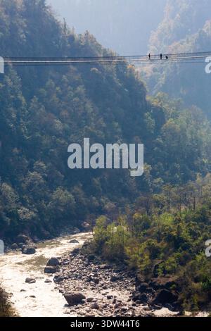 Drei Menschen überqueren eine hohe Hängebrücke über einen malerischen Fluss in Nepal. Stockfoto