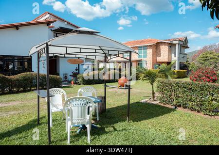 Geräumiger Wohngarten mit einem weißen Gartenlaube mit Kunststoff-Terrassenmöbeln, bietet einen komfortablen, schattigen Bereich inmitten üppiger Hecken und Stockfoto