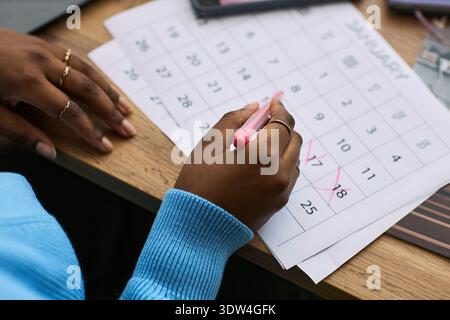 Schwarze Frau markiert Datumsangaben auf dem Kalender mit rosa Stift, fokussiert auf bestimmte Tage im Januar, Gesundheit Tracking Konzept, Nahaufnahme der Hände und Kalender Stockfoto