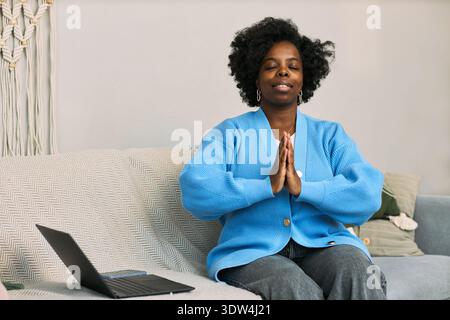 Schwarze Frau junger Erwachsener sitzt auf Sofa und übt Achtsamkeitsmeditation mit Augen geschlossenen Händen in Gebetsposition, Laptop auf Sofa fördert Gesundheit und Wellness Stockfoto