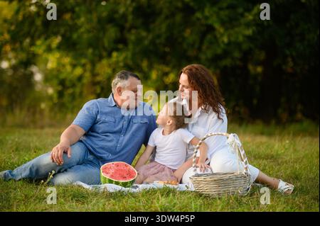 Die Familie genießt ein Picknick im Park, sitzt zusammen auf dem Gras, lächelt und plaudert. Mom, Dad, Tochter verbringen Zeit mit fru im Freien Stockfoto