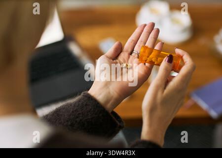 Kaukasische Frau mittleren Alters, die verschreibungspflichtige Pillen in der Hand hält und Medikamente in die Hand gießt, mit Laptop und Notebook am Schreibtisch sitzt und das Frauengesundheitsmanagement veranschaulicht Stockfoto
