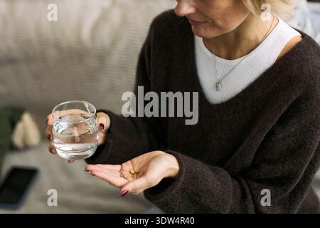 Weiße Frau mittleren Alters, die Glas Wasser in der einen Hand und mehrere Pillen in der anderen Hand hält, sich auf Medikamente vorbereiten, drinnen sitzen, sich auf Gesundheit und Wellness konzentrieren Stockfoto