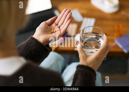 Kaukasische reife Frau, die ein Glas Wasser in der einen Hand und mehrere Pillen in der anderen Hand hält, am Schreibtisch sitzt mit Medikamentenblase und Laptop im Hintergrund, Schwerpunkt auf Gesundheit Stockfoto