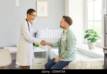 Besprechung des Klinikraums, Ärztin schüttelt die Hände mit männlichem Patienten, der auf dem Untersuchungsbett sitzt Stockfoto