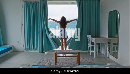 Die Leute entspannen sich im Bali Resort. Die Frau steht im Hotelzimmer und zieht die Vorhänge aus blauem Grün zurück, um die wunderschöne Berglandschaft über dem Mount Batur und dem See zu enthüllen. Sie fühlt Urlaubs- und Reisemoment. Rückansicht Stockfoto