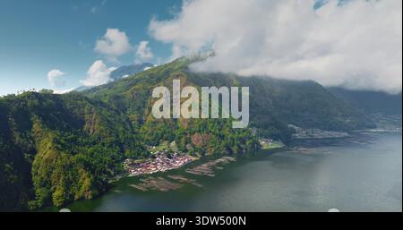 Indonesien, Bali, Abang Dorf und traditionelle Aquakulturflöße, die auf dem ruhigen Wasser des Lake Batur schwimmen, grüne Hügel und Wolken über den Gipfeln des Vulkans Mount Abang. Flugpanorama der Drohne Stockfoto