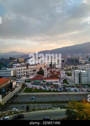 Erhöhter Panoramablick auf den zentralen Stadtplatz mit Denkmal, historischen Gebäuden, belebten Straßen und Bergen unter warmem Abendhimmel. Stockfoto