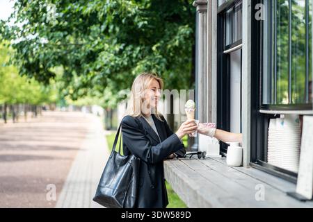 Glückliche, fröhliche Geschäftsfrau, die nach einem anstrengenden Arbeitstag Eis am Straßenkiosk im Stadtpark kaufte. Stockfoto