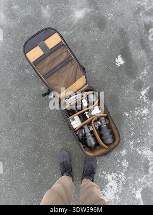 Top-Ansicht einer offenen professionellen Kameratasche auf dem gefrorenen See mit spiegellosen Kameras, Objektiven, Drohnen, Batterien und Füßen des Fotografen in Sneakers. B Stockfoto