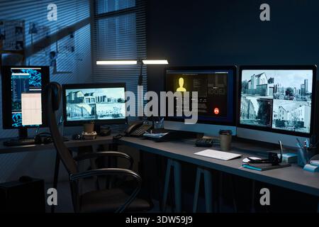 Modernes Überwachungsbüro mit mehreren Computermonitoren, die Überwachungskameras von Gebäuden und digitalen Profilen anzeigen, leeren Arbeitsplätzen mit elektronischen Geräten und Zubehör Stockfoto