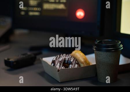 Nahaufnahme mit Schokoladengarnitur mit Glasur und Einweg-Kaffeetasse auf dem Schreibtisch in der Nähe von Computermonitoren und Walkie-Talkie, die einen Snack am Arbeitsplatz oder eine Nachtschicht-Pausenszene veranschaulicht Stockfoto