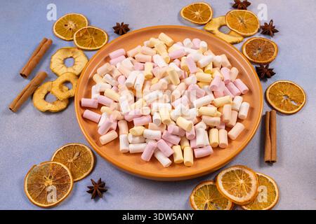 Farbenfrohe Mini-Marshmallows, die einen orangefarbenen Teller füllen, umgeben von verstreuten getrockneten Orangenscheiben, Zimtstäbchen und Sternanis auf einem hellblauen Hintergrund Stockfoto