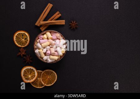 Kleine bunte Marshmallows, die eine Schüssel füllen, angeordnet mit getrockneten Orangenscheiben, Zimtstangen und Sternanis auf dunklem Hintergrund, schaffen ein gemütliches W Stockfoto