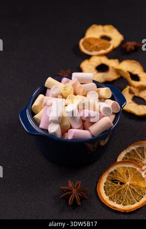 Bunte Marshmallows in einer blauen Schüssel, bereit für heiße Schokolade oder Kakao, umgeben von getrockneten Orangen, getrockneten Apfelscheiben und Sternanis auf einem dunklen Text Stockfoto