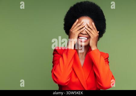 Aufgeregt staunte afrikanische Frau bedeckt Augen emotional von einem angenehmen überraschenden Geschäftssieg oder Lotteriegewinn. Elegante afro-amerikanische Geschäftsfrau in rotem Anzug, überglücklich mit guten Nachrichten, isolierter Studioaufnahme || Model veröffentlicht Stockfoto