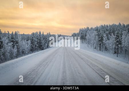 Blick auf eine lange Straße, die sich durch einen schneebedeckten Wald unter einem weichen, warmen Himmel erstreckt und eine ruhige Winterlandschaft schafft, Ruka, Pohjois-Pohjanmaa, Finl Stockfoto