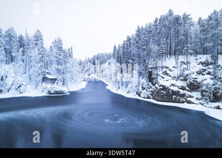 Blick auf den wirbelnden, eisigen Fluss, der sich durch einen schneebedeckten Wald schlängelt, eine kleine Hütte auf dem schneebedeckten Ufer in Ruka, Pohjois-Pohjanmaa, Finnland. Stockfoto