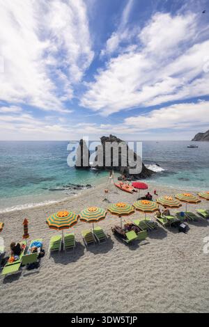 Blick auf den sonnenverwöhnten Strand mit farbenfrohen Sonnenschirmen, die sich vom türkisfarbenen Meer abheben, eingerahmt von zerklüfteten Klippen in Monterosso al Mare, Ligurien, Stockfoto