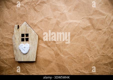 Hölzerne Hausfigur mit weißem gestricktem Herzen auf zerknittertem Kraftpapier-Hintergrund. Das Konzept der häuslichen Liebe, des Familienkomforts, der Immobilien, der Hypothek, der Versicherung Stockfoto