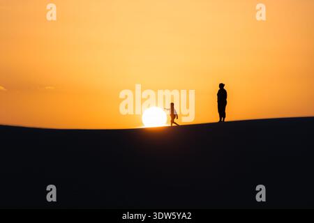 Blick auf die Silhouetten von Menschen auf einem Kamm vor einem leuchtend orangefarbenen Sonnenuntergangshimmel, der lange Schatten in da'ira, Socotra, Jemen wirft. Stockfoto