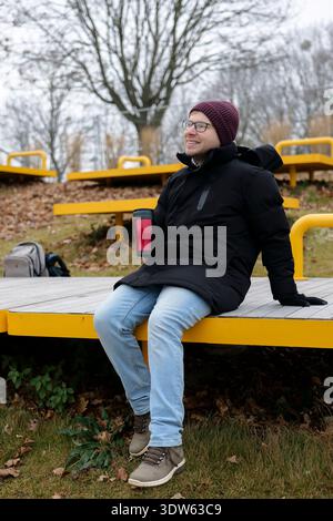 Junger lächelnder Mann, der im Winter ein warmes Getränk aus einem roten Reisebecher auf einer gelben Bank genießt. Konzept einer städtischen Winteraktivität Stockfoto