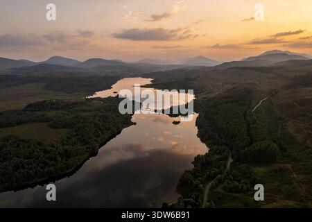 Ein Panorama-Drohnenfoto fängt das goldene Spiegelbild der untergehenden Sonne auf einem gewundenen See ein, der von grünen Wäldern und Bergen umgeben ist. Stockfoto