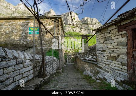 Blick auf einen schmalen Steinweg, der sich zwischen alten Gebäuden schlängelt und den Blick in Richtung der majestätischen Berge, Mikro Papigko, Ioannina, Griechenland, führt. Stockfoto