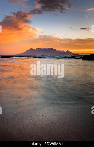 Blick auf den legendären Tafelberg vor einem feurigen Sonnenuntergang, der den Himmel mit leuchtenden Orangen und Blautönen übermalt und sich im nassen Sand, CA, spiegelt Stockfoto