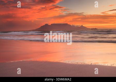 Blick auf den legendären Tafelberg, der sich vor einem feurigen Sonnenunterganghimmel befindet, Wellen, die sanft das Sandstrand wäschen und eine ruhige Landschaft schaffen, Kapstadt Stockfoto