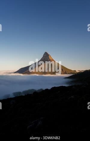 Der Blick auf einen einsamen Lion's Head Berg erhebt sich majestätisch über einem Meer ätherischer Wolken bei Sonnenuntergang, das goldene Licht wirft lange Schatten, Cape Stockfoto