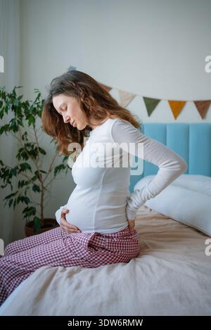 Schwangere Frau, die unter Schmerzen im unteren Rücken leidet und die eine Hand auf den schmerzenden Rücken und die andere auf den Bauch des Babys legt, was die körperlichen Herausforderungen und Beschwerden der Mutterschaft widerspiegelt Stockfoto
