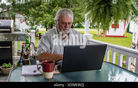 Ein Mann sitzt an einem Tisch auf einer Veranda und arbeitet an einem Laptop, während er Kaffee trinkt. Er hat weiße Gesichtshaare und trägt ein helles Hemd. Der Hinterhof ist grün mit Bäumen und einem Grill in der Nähe. Stockfoto