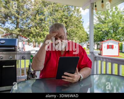 Ein Mann sitzt an einem Tisch auf einer Veranda und schaut auf eine Tafel. Er ist draußen in einem Hinterhof mit Bäumen und einem Grill in der Nähe. Es ist ein heller, sonniger Tag mit klarem Himmel. Stockfoto