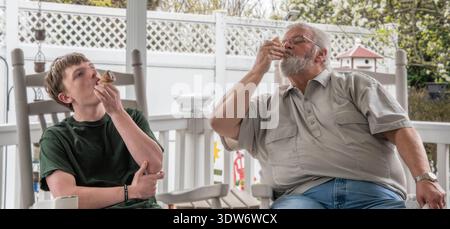 Ein junger Mann und ein älterer Mann sitzen auf einer Veranda und essen Snacks. Sie haben ein Lächeln im Gesicht, während sie ihre gemeinsame Zeit genießen. Der Hintergrund zeigt einen grünen Garten und einen sonnigen Tag. Stockfoto